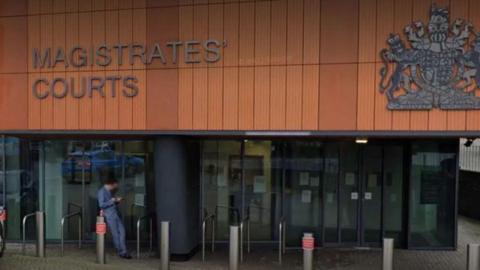 Close-up of Colchester Magistrates' Court's entrance. There are seven metal bollards outside the automatic doors. A person in a light blue suit is stood to the left of the image and is looking down at his phone. 