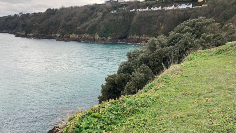 An expanse of sea with part of the island seen in the background, with rocks in the bottom of the photo. Grey and blue skies.