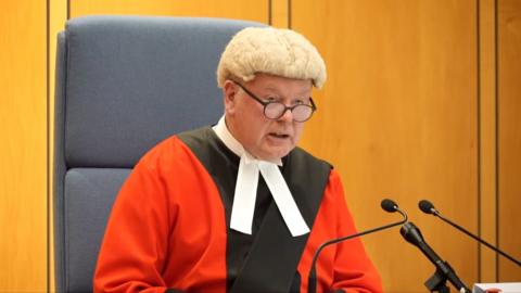 Judge in white wig and a red and black gown addresses Neil Trennan who is off frame.