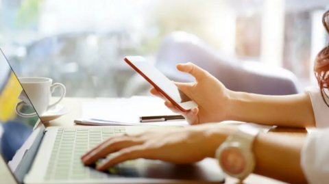 A woman uses a laptop while holding a mobile phone in her right hand.