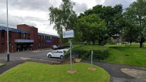 The front of a police station. In the foreground are grass verges, a sign indicating the station and parked cars. The station itself is a red-brick building set back from the road.