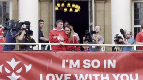Craig Eskrett, wearing a red jumper, stood with a woman, who is also wearing a red jumper, and a child on City Hall's balcony. Media crews with cameras are surrounding them. A large red banner reads 'I’M SO IN LOVE WITH YOU' in capitals with white logos on both sides.