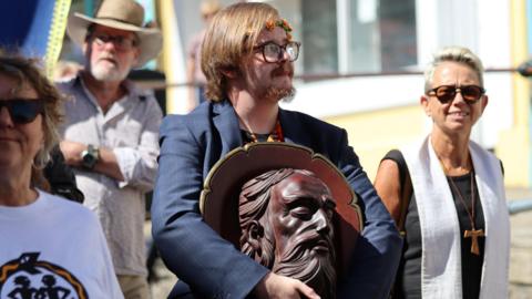 A man with a ring of orange flowers in his hair carries a wood head of St John during a parade. He is wearing a blue suit jacket. He also has glasses on and has straight brown hair and a brown goatee beard. Two women are stood either side of him and a man wearing a hat is behind him.