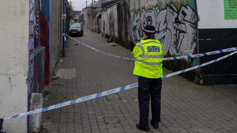 A police office wearing a yellow hi-vis jacket stood in front of blue and white police tape. The tape is covering an alley way with graffiti on the walls. 