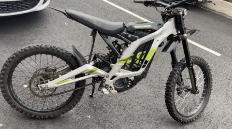 An electric bike on its stand in a car park. It has a white and yellow livery and the words "SUR RON" printed near the seat. The wheels have a thick tread. 