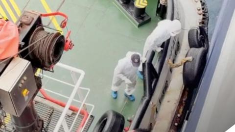 People in hazmat suits on a boat seen from above - they wear blue protective shoe coverings and one is looking over the edge of the vessel 