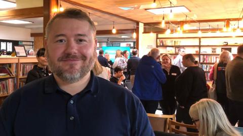 Ian Sharp smiles as he stands in a well-lit room during a busy networking event. He has short, light-brown hair and a matching, neatly trimmed beard. He wears a blue polo shirt. Groups of people can be seen talking in the background.