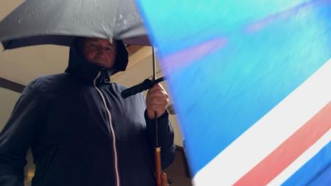 Stephen Harris, attempting to hide using umbrellas held by himself and another man who accompanied him out of court. The other umbrella bears a Union Flag pattern.