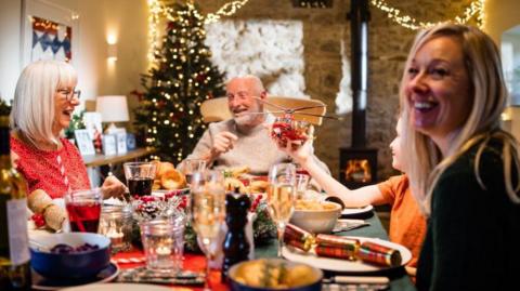 A family sit around the dinner table at Christmas. They are smiling and laughing. The child is holding a toy air ambulance.
