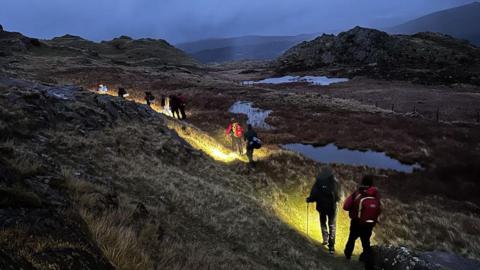 A line of people walking in pairs away into the distance on misty Lake District fells. They are using torches to light their way across rough grassland dotted with pools of water. The landscape is rocky and more mountains can be seen in the gloomy distance.