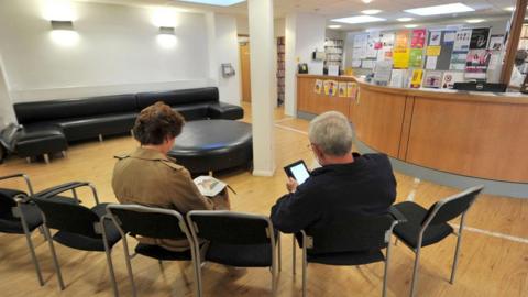 A woman and a man sitting on chairs in the waiting room of a GP Practice, with the reception desk in the background