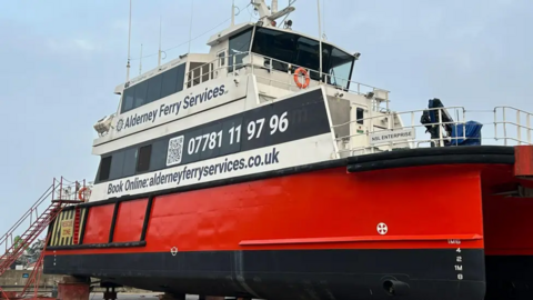 The Dart Fisher ferry boat out of the water, with a red hull and white upper deck displaying Alderney Ferry Services contact details