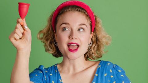 Comedian Bella Humphries wearing a red headband with mouth slightly open  holding a mooncup to the left and looking up towards it. 