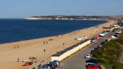 A shot over Seaford beach on a sunny day. There are a number of people along the long stretch of coast with a white cliff in the background.