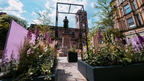 Plants and flowers line both sides of an arch over a narrow path in St Ann's Square. The old two-storey church with its clock tower is in the background with a statue in front seen between the arch.