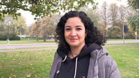 Helaleh Cheraghmakani is pictured in a grey coat and black hoodie. in the background is green grass and trees.