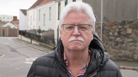 Chris Band: A man in his 60s wearing a black puffer jacket and a blue shirt with a red‑and‑white striped collar is standing in the middle of a quiet street. The background shows a residential area with light‑coloured buildings, a stone wall, and a narrow road curving to the left. The sky appears overcast.