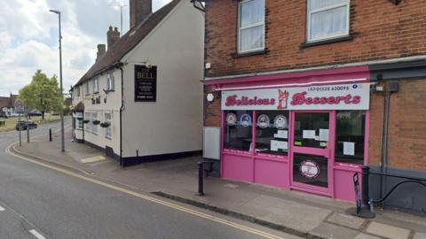 The pink shop front of Delicious Desserts, which is housed in a red brick two storey building. The sign is in bubble writing and has an image of an ice cream sundae. It is next to the Bell pub and near the village green. 