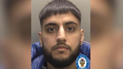Mohammed Ibrahim in a mugshot. He has dark hair and beard, and a neutral expression on his face. He is wearing a blue puffer jacket.