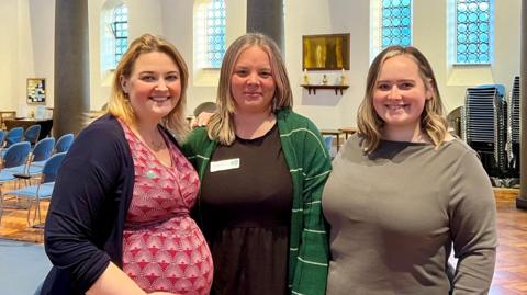 Three women smile at camera. The woman on the left has a pink dress on, the woman in the middle has a black dress, and the woman on the right has a grey top on. They stand in a hall with a wooden floor and several blue chairs can be seen behind them. A metal plaque is on the wall behind them with three candlestick holders on a shelf underneath.