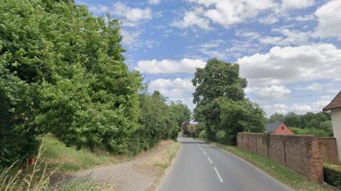 The Street in Monks Eleigh. The road is lined by small cottages, tall trees and leafy bushes. It is a 30 miles per hour limit and is slightly bendy.