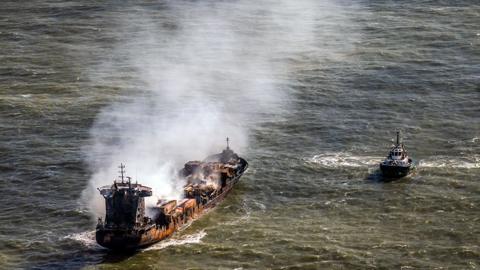 The Solong being shadowed by another ship. Smoke is coming out of the vessel and the hull and superstructure has burn marks across it