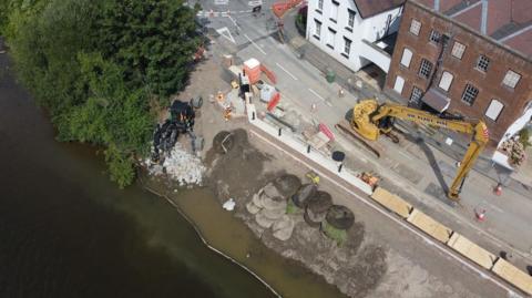A large yellow machine is in the road near where the wall was being built by trees and the river.