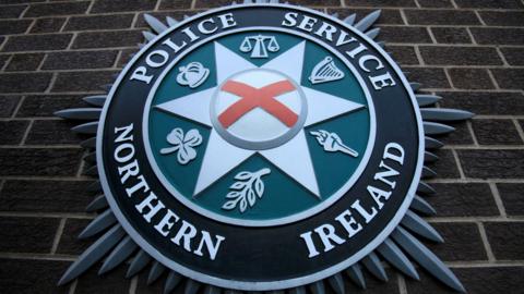 A close-up of the PSNI crest on brown brick wall.  The badge has a red cross in the centre of a star, surrounded by six emblems with include a harp, a crown, a shamrock and the scales of justice.  The centre of the badge is surrounded by a black circle featuring the words Police Service of Northern Ireland in capital letters. 