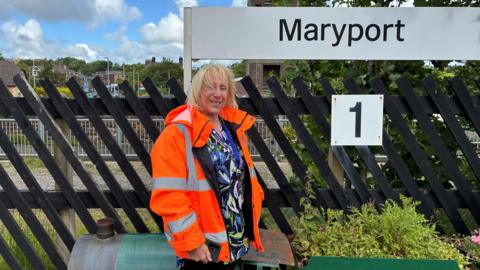 Jane Murray wearing a bright orange high-visibility jacket stands in front of a brown wooden fence at Maryport railway station. A large white sign reading Maryport is mounted above, and a smaller sign with the number 1 indicates the platform. Behind the fence are train tracks, greenery, and buildings under a partly cloudy sky.