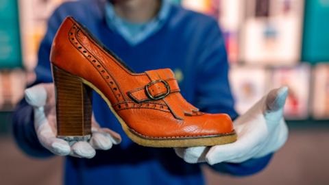 An elegant high-heeled shoe is held in the hands of a member of staff at the Clarks museum in Somerset. He is wearing white gloves. The shoe has a thick heel and intricate stitching and is light brown in colour