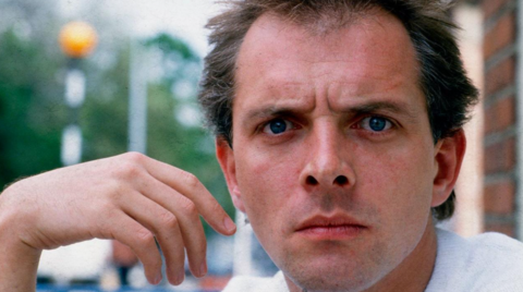 Mayall, a clean-shaven man with dark hair, looks at the camera. His hand is raised and hangs limp by the side of his face. The background is blurred.