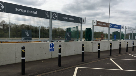 A household waste recycling centre with signs for scrap metal, wood and timber and cardboard. In front of the signs there are concrete barriers and a line of bollards.