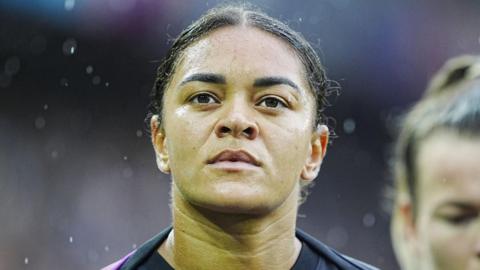 Jess Carter, who has her dark hair in a ponytail, wearing a warm-up top ahead of a football match on a rainy day.