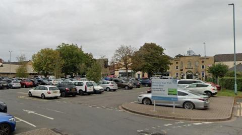 A full car park in Cirencester, with trees and buildings in the background on a cloudy day.