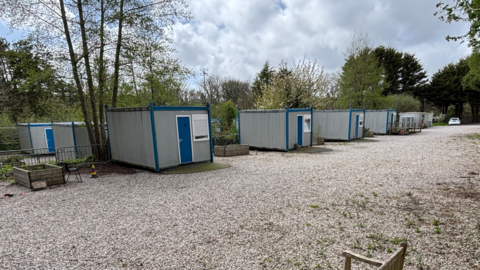 The picture shows a small cluster of portable cabin units set out on a gravel surface in a quiet, leafy area. The cabins are rectangular, grey in colour, with blue trim and blue doors, and they appear to be arranged in a loose line running away from the camera. Each unit looks self contained, with small windows, and some have raised planters or barriers placed nearby.