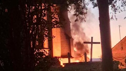 Churchyard with orange glow of fire and smoke billowing into the sky. A number of crosses can be seen in the cemetery.