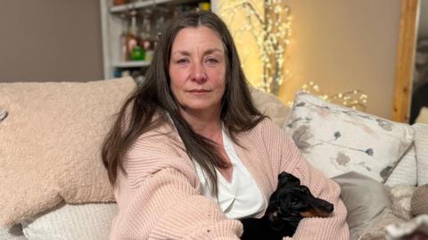 Image shows a lady named Rebecca Rumens sat on a sofa. She is wearing a pink knitted cardigan and a white top. She has black trousers on. She has long hair that is parted down the middle. On her lap is a black dog sleeping. Behind her are a selection of cushions which are blush pink coloured and beige. There is a mirror to the right of the picture. There are fairy lights the background.
