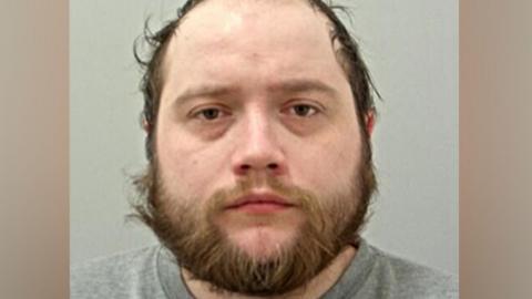 Custody photo of Dylan Saville wearing a grey sweatshirt while against a light grey background. He has lots of brown facial hair and brown eyes. 