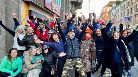 A group of 20 to 30 people standing with their arms up and smiling outside the YMCA. One man in the centre is wearing aviator sunglasses, camo trousers, a dark blue gilet and blue top, with a pilot's hat on.