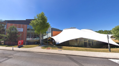 A screenshot from Google street view showing a building with a large white roof. There's a sight next to the building with the red Vodafone logo on it and text reading "hello". It's a sunny day and the sky is blue.
