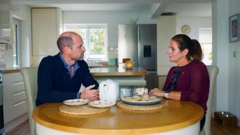 Prince Williams and Rhian Mannings sat around her kitchen table talking with a cup of tea. On the table there is a tea pot, two heart patterned plates and a plate of Welsh cakes. Williams, on the left, wears a navy quarter zip and a light blue shirt. On the right, Rhian wears a long sleeved burgundy blouse with black bow details. She has dark brown hair in a ponytail. 