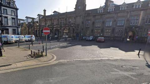 The square outside Carlisle railway station station, an ornate, long three-storey building with cars and vans parked outside.