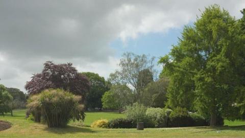 Courtenay Park, Newton Abbot. A large green park. There are trees and bushes and green grass. The sky is light blue and cloudy. 