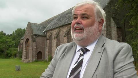 Torbay Council leader David Thomas stood in front of the Spanish Barn in Torquay. Mr Thomas has white hair and a white beard and is wearing a grey blazer, white shirt and a black, white and grey striped tie. He has his mouth slightly open mid-speech. The Spanish Barn is a windowless stone building with a large wooden door.