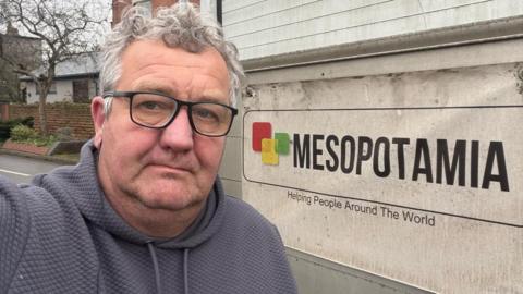 Richard Newbold, in a grey hoodie, looking at the camera, stood next to a van with a Mesopotamia banner on it