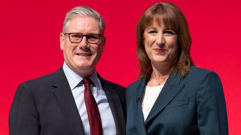 Starmer and Reeves in front of a bright red background smiling at the camera.