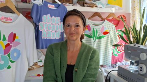 Amy Laws looks at the camera. She is sitting down with a sewing machine to the right and lots of T-shirts around her - all have been adapted with bold, colourful designs, mostly floral. She is wearing a green cardigan and a black t-shirt and headband