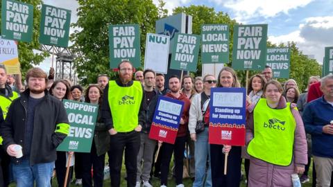 A crowd of protesters stand on grass holding signs reading “Fair Pay Now”, “Support Local News” and “Standing up for Journalism”. Several people wear bright yellow Bectu union bibs, while others hold NUJ placards during the demonstration under a cloudy sky.