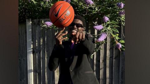 Leon Pafu Ngoy is smiling broadly while holding a basketball and pointing his finger at the camera. He is wearing a black suit and sunglasses and is standing in front of metal railings, with a plant with purple flowers behind him.