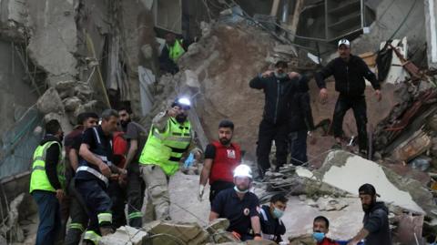 A number of rescue workers, some with helmets and head torches, others with face masks, work in the rubble of a destroyed building